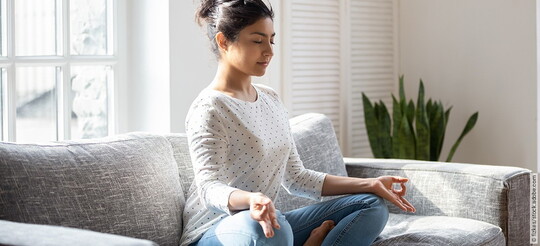 Junge Frau meditiert auf dem Sofa, um die Technik zur Selbsthilfe bei Tinnitus einzusetzen.