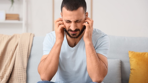 Mann auf der Couch hält sich die Ohren: Er leidet unter subjektivem Tinnitus – das ist nur eine von verschiedenen Tinnitus-Arten.