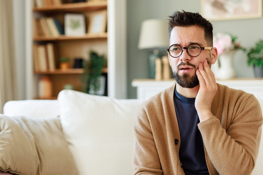 Ohrenerkrankungen wie Mittelohrentzündungen als Ursache für Tinnitus: Mann sitzt auf Sofa und hält Hand an sein schmerzendes Kiefergelenk.